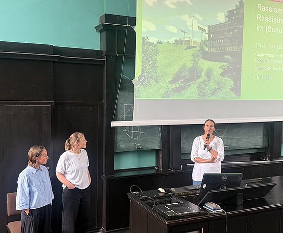 Rednerin steht mit einem Mikrofon in der Hand vor einem Pult in einem Hörsaal, im Hintergrund eine Präsentationsfolie auf einer Leinwand. Tina Nobis und Lara Kronenbitter stehen links davon und hören zu.