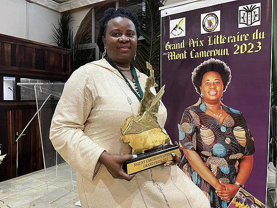 Priscillia Manjoh sitzt vor einem Plakat mit einem großen Foto von sich und hält Trophäe in den Händen