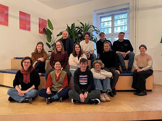 Projektgruppe sitzt und steht auf einer mehrstufigen Holzplattform vor einer Wand mit drei roten quadratischen Bildern und einer großen Zimmerpflanze neben einem Fenster