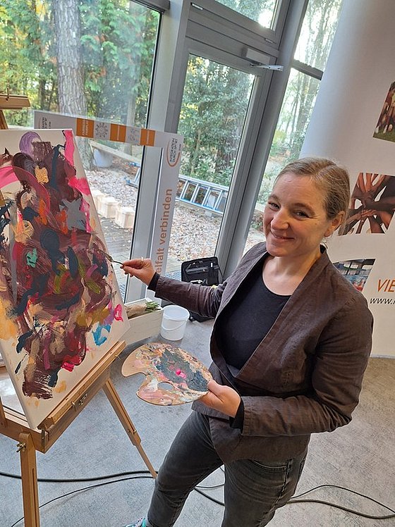 Tina Nobis steht vor Staffelei und malt mit Pinsel auf Leinwand, hält eine Farbpalette in der linken Hand