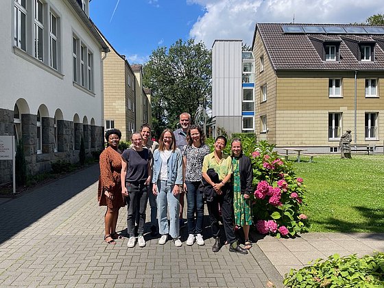 Team der Sportsoziologie steht auf einem gepflasterten Weg zwischen zwei Gebäuden bei sonnigem Himmel